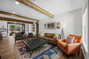 A modern living room with a sofa, ottoman, and chairs, a patterned rug, and a kitchen in the background.