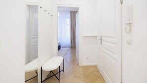 Een smalle gang met witte muren, een bankje en een deur naar een kamer met een bed en gordijnen.