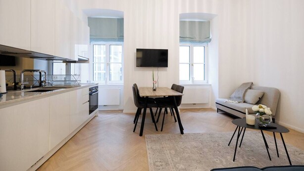 Een moderne keuken met witte kastjes, een eethoek met tafel en stoelen, een bank en een aan de muur bevestigde televisie.