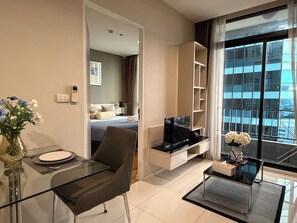 A modern hotel room with a glass dining table, a comfortable chair, a flat-screen TV, a small side table, and a large window with a city view. - Balcony & Kitchen · Pool, Gym, Fast Wi-Fi (Watthana)