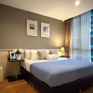 A modern hotel room with a large bed, a nightstand with a lamp, and two framed pictures on the wall. - Balcony & Kitchen · Pool, Gym, Fast Wi-Fi (Watthana)