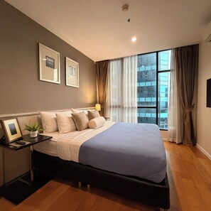 A modern bedroom with a large bed, a bedside table with a plant, and a window with curtains. - Balcony & Kitchen · Pool, Gym, Fast Wi-Fi (Watthana)