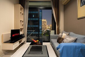 A modern living room with a Netflix TV, a glass coffee table with fruit, a grey sofa, and a city view through a large window. - Balcony & Kitchen · Pool, Gym, Fast Wi-Fi (Watthana)