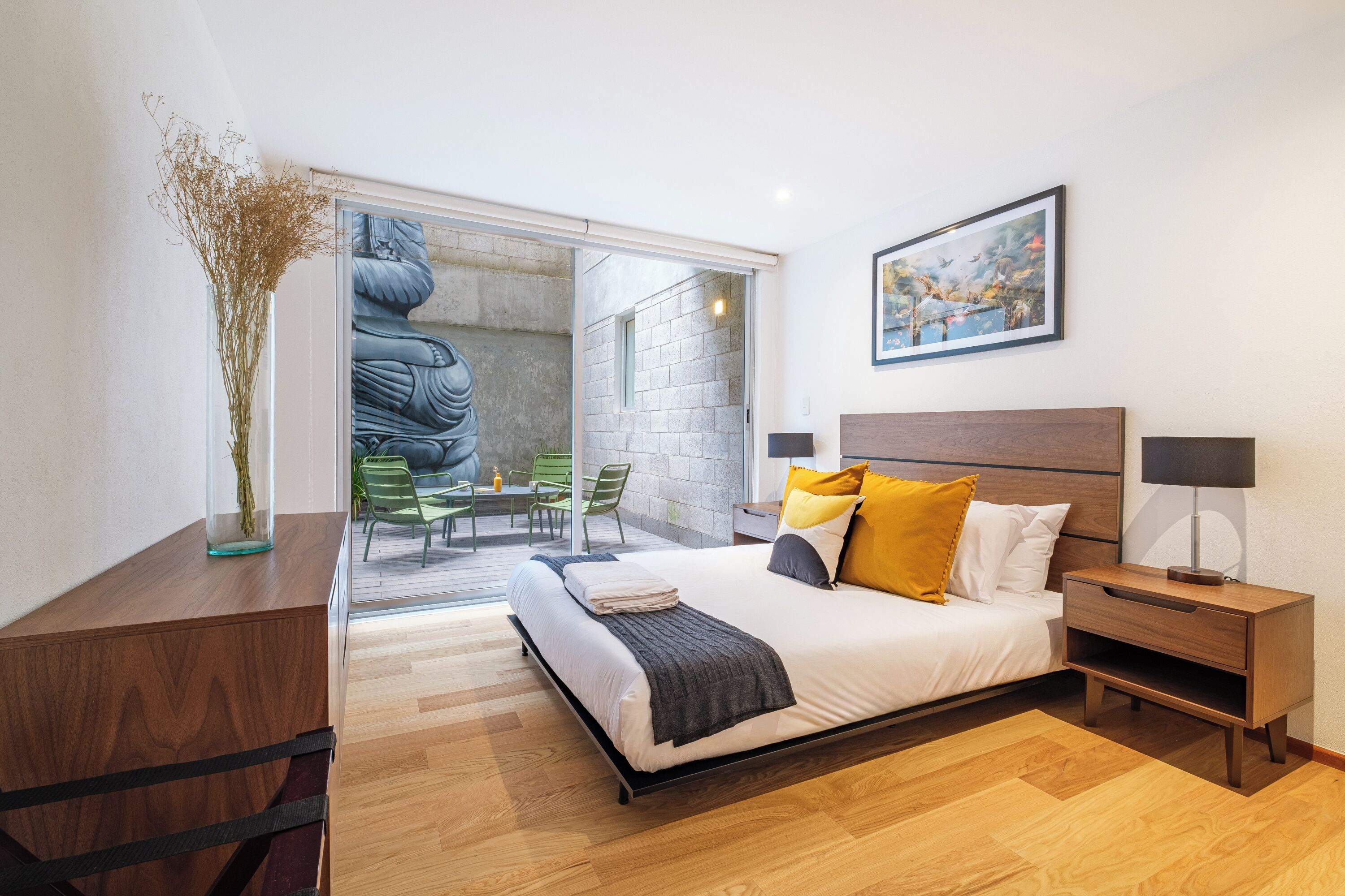 Ein modernes Schlafzimmer mit einem Bett, Nachttischen, einer Glastür, die auf einen Balkon führt, und einem gerahmten Bild an der Wand.