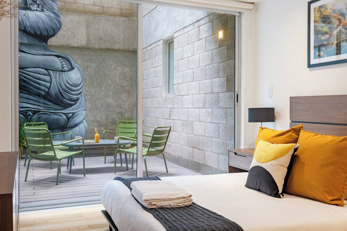 Ein Schlafzimmer mit Bett, Nachttisch, Lampe, Fenster und Blick auf eine Steinmauer und eine Statue.