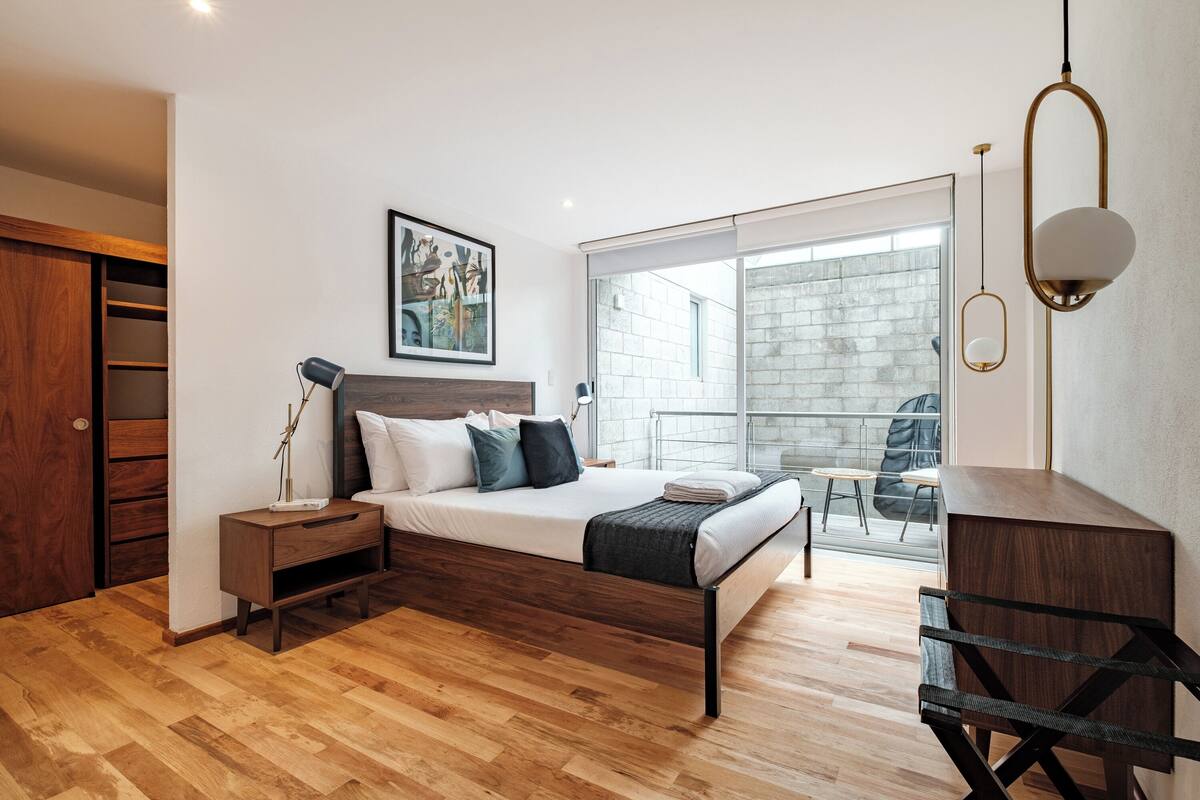 Ein modernes Schlafzimmer mit einem hölzernen Bett, einem Nachttisch, einem Schreibtisch und einem Balkon mit Blick auf ein Backsteingebäude.