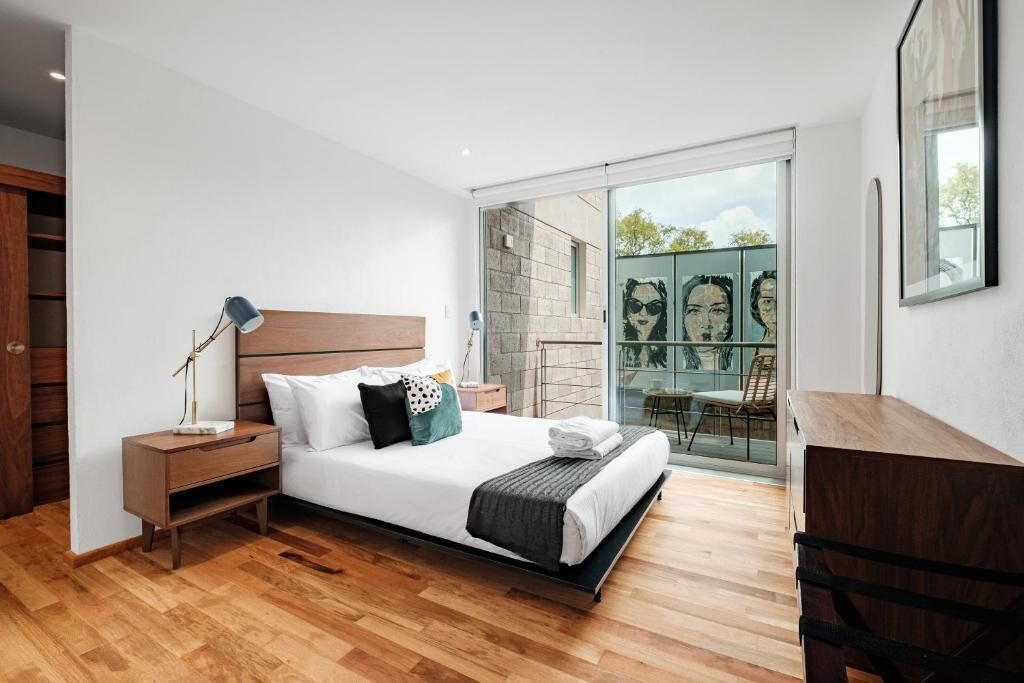 Ein modernes Schlafzimmer mit Holzboden, einem Bett mit weißer Bettwäsche, einem Nachttisch, einem Schreibtisch und einer Glastür, die auf einen Balkon mit Sitzgelegenheit führt.