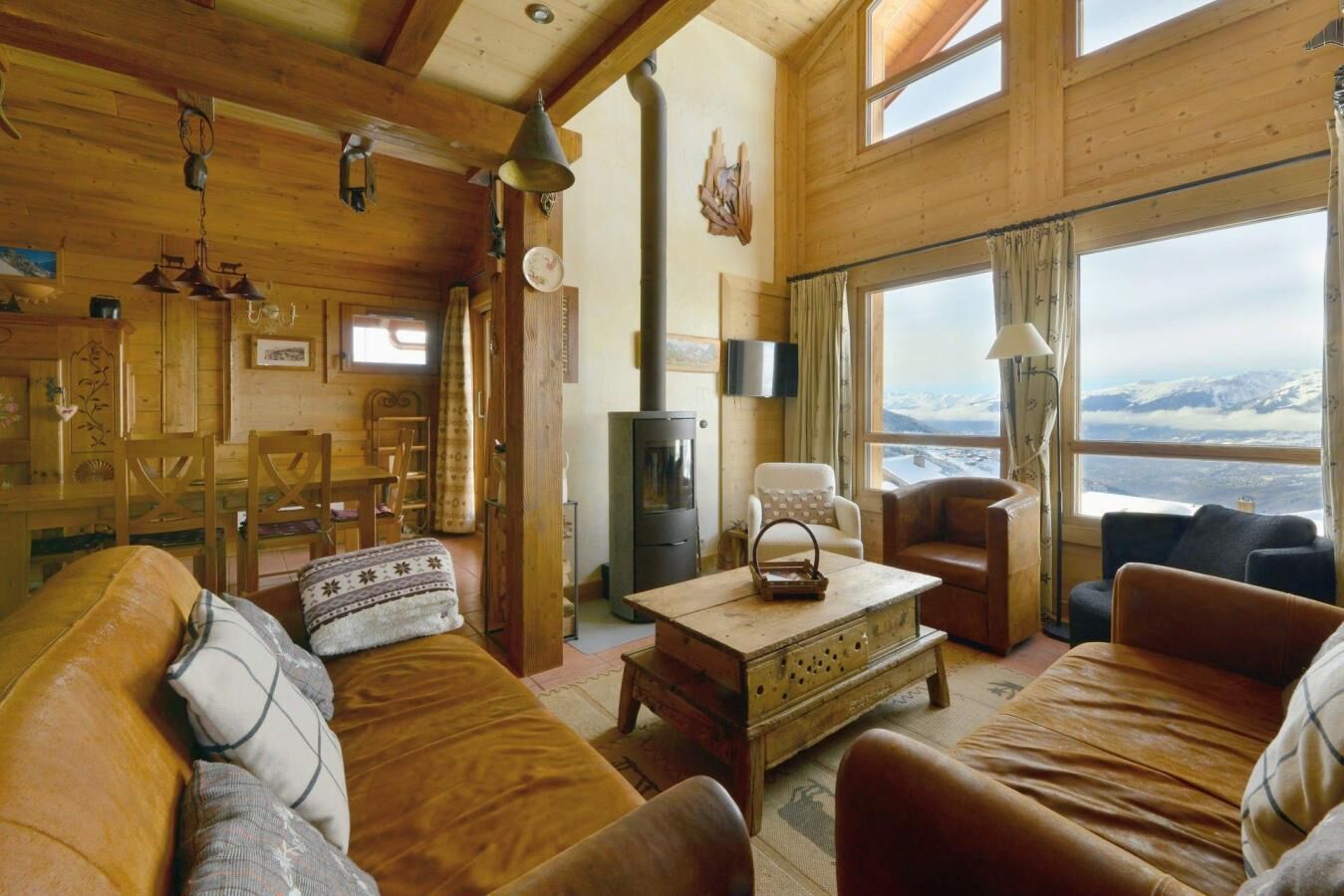 A cozy cabin interior with wooden walls, a fireplace, a dining area, and a large window offering a mountain view.