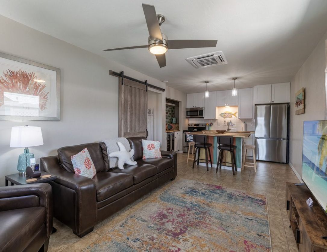 A modern living room with a sofa, a coffee table, and a TV. There is a kitchen area with a refrigerator, microwave, and a dining table.