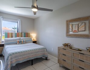 Ein Schlafzimmer mit Bett, Deckenventilator, Nachttisch mit Lampe, Kommode und einem Fenster mit Vorhängen.