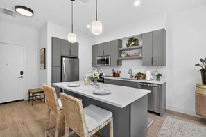 Eine moderne Küche mit grauen Schränken, einem Edelstahlkühlschrank, einer Mikrowelle und einem Geschirrspüler. Eine weiße Kücheninsel bietet Platz für zwei Stühle und eine Blumenvase.