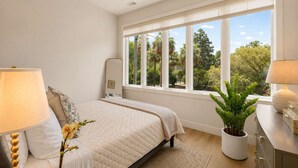 Un dormitorio con cama, lámparas de noche, un ventanal con vistas a árboles, un ventilador de techo y una planta en maceta.