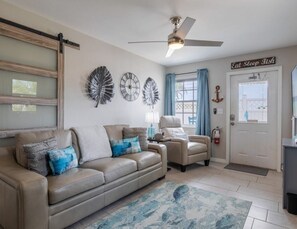 A living room with a sofa, armchairs, a ceiling fan, decorative wall art, and a door with a sign. - 1 bedroom apartment in Saint James City with AC -Tarpon Suite (Saint James City)