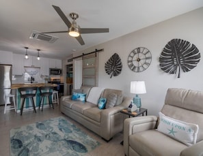Ein modernes Wohnzimmer mit Sofa, Sessel und Couchtisch. Es gibt eine Küchenzeile mit einer Bar und Hockern. Dekorative Wanduhren und ein Deckenventilator sind zu sehen.