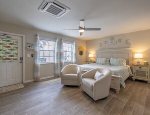 Ein Schlafzimmer mit einem Bett, zwei Stühlen, einem Nachttisch, einer Lampe, einem Deckenventilator und einem Fenster mit Vorhängen.
