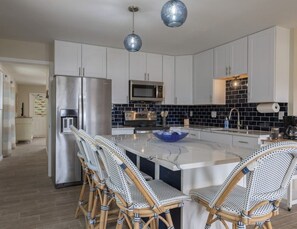 Eine moderne Küche mit weißen Schränken, einem Edelstahl-Kühlschrank, einer Mikrowelle und einer schwarzen Küchenzeile aus Fliesen. Körbchenstühle umgeben eine zentrale Kochinsel mit einer blauen Schüssel.
