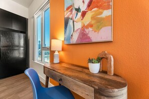 Una habitación con un escritorio de madera, una silla azul, una lámpara, una planta en maceta y un cuadro abstracto y colorido en la pared.
