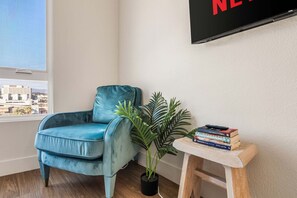 A room with a blue armchair, a potted plant, a small wooden table with books, and a TV mounted on the wall. - Stylish Haven with Scenic DTLA Views & Parking - Walking to Little Tokyo (Los Angeles)