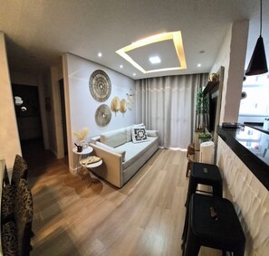 A modern living room with a sofa, decorative wall art, a small table with a vase, and a black stool. - Premium Apartment with Garage | Near Riocentro and Olympic Park (Rio de Janeiro)