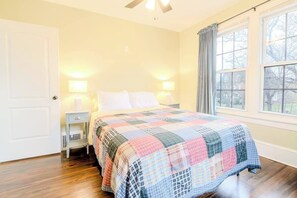 Ein Schlafzimmer mit einem Bett, Nachttischlampen, einem Beistelltisch, einem Fenster mit Vorhängen und einem Deckenventilator.