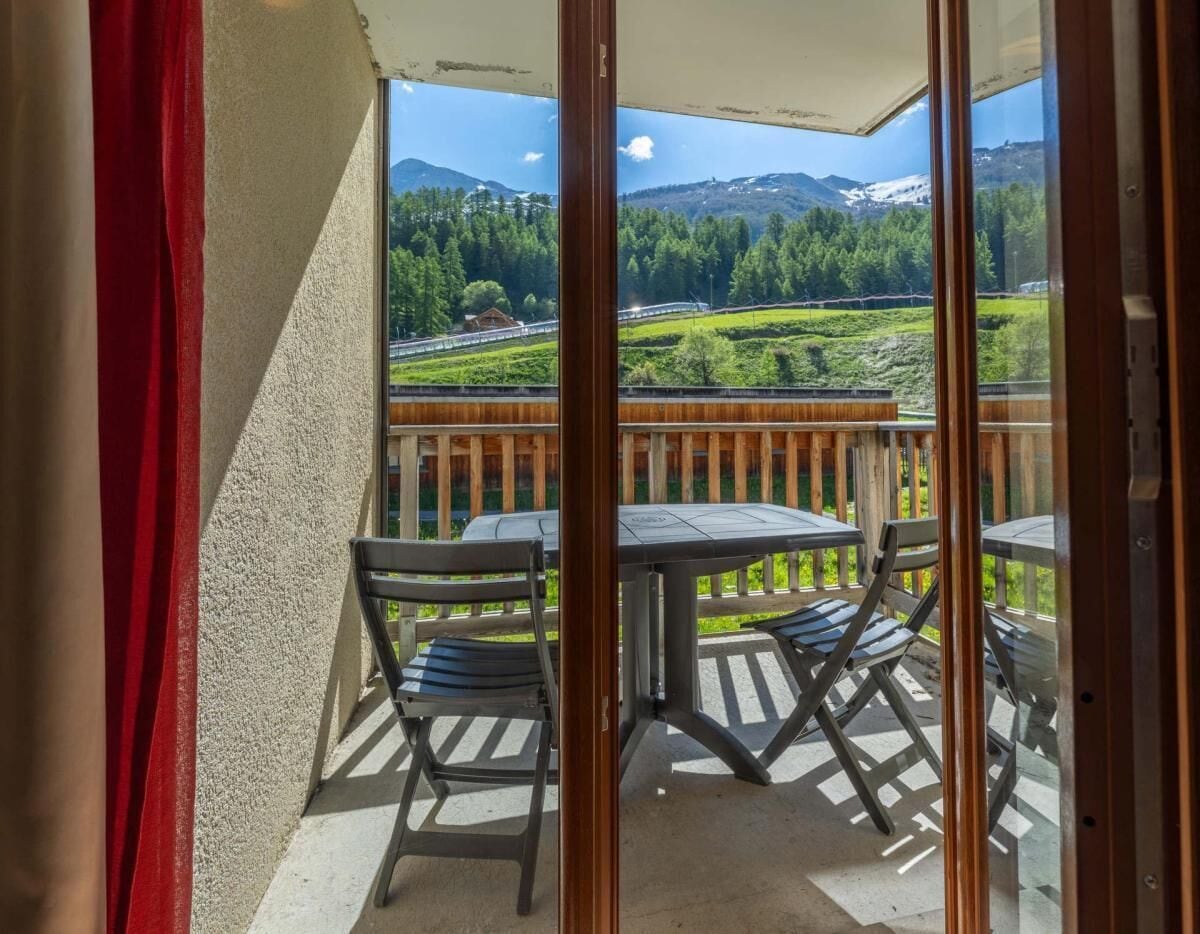 A balcony with a table and chairs, a wooden railing, and a view of a green landscape with mountains.
