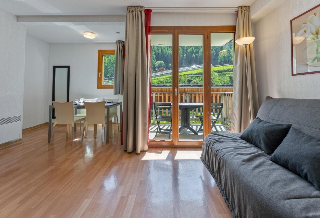 Une pièce dotée d’un plancher en bois, d’un canapé, d’une table à manger avec des chaises et d’une porte-fenêtre coulissante donnant sur un espace extérieur avec vue sur la verdure et une route.