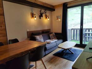 A cozy room with a wooden dining table, a grey sofa with pillows, a wooden bench, and a large window with a view of greenery. - 40m from ski slopes, Ground floor, fitness, swimming pool, sauna, balcony, tv, ski locker, 35m² (Les Orres)