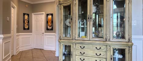 A hallway with a large, ornate cabinet with multiple drawers and glass doors, white wainscoting, and a tiled floor.