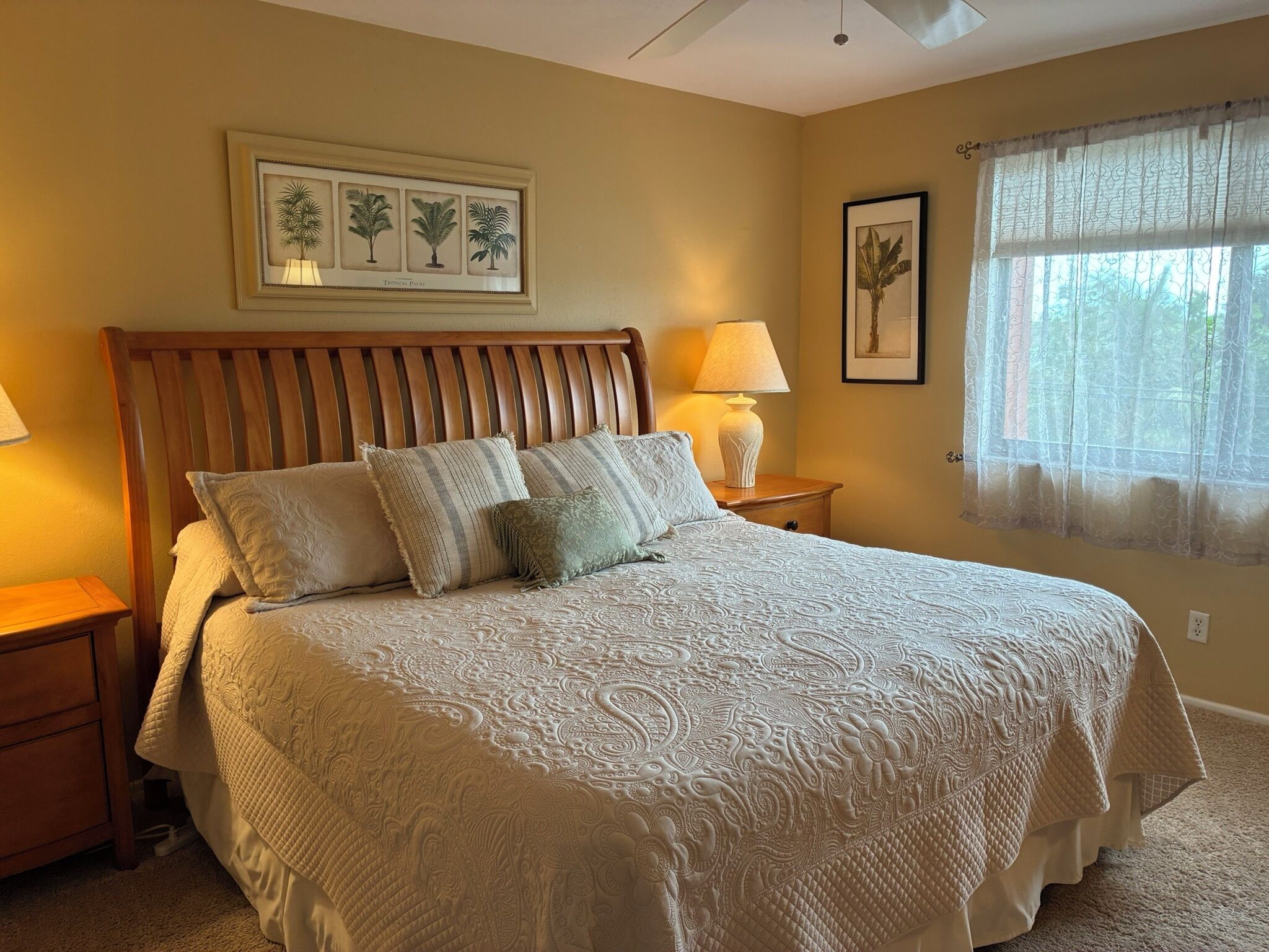A bedroom with a large bed, a wooden headboard, two bedside tables, a wall-mounted lamp, a framed botanical print, and a window with sheer curtains.