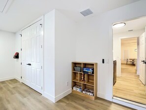 Un couloir avec une porte blanche, une petite étagère en bois avec des boîtes, et une vue sur une autre pièce qui comprend un coin repas.