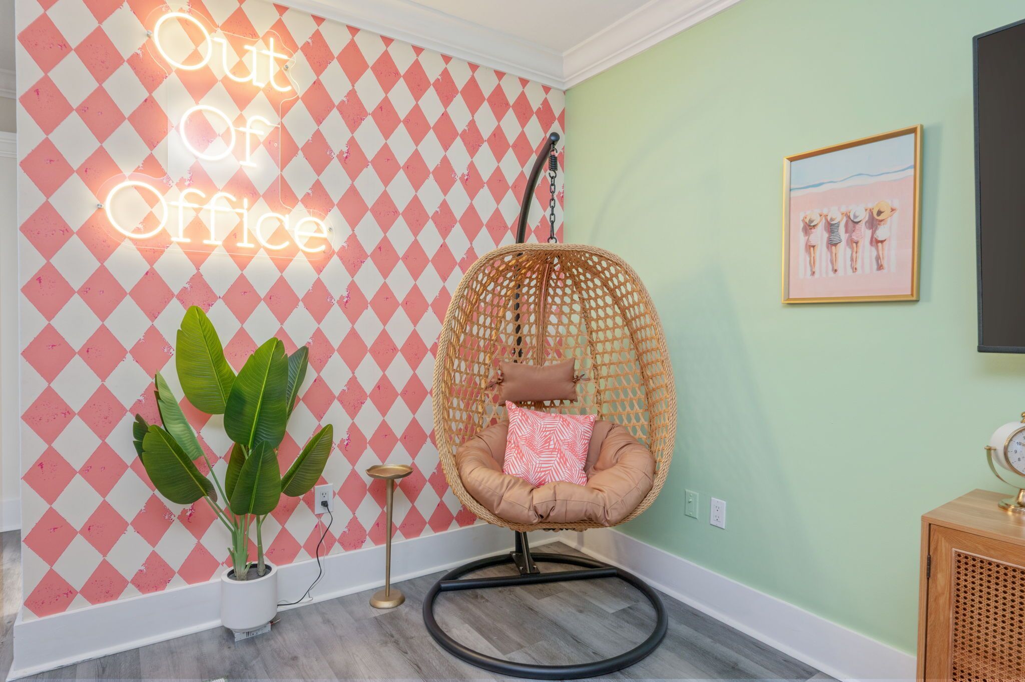 A room with a neon sign reading 'Out of Office,' a wicker chair with a cushion, a potted plant, and a framed picture on the wall.