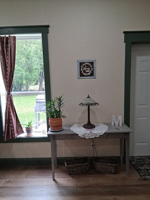 Una habitación con ventana, una mesa con lámpara, una planta en maceta, un cartel enmarcado y una puerta.