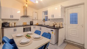 A modern kitchen with white cabinets, a dining table with blue chairs, a built-in oven, and a washing machine. - Crème Da La Casa (Coventry)