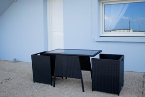 Une table en osier noir, accompagnée de deux chaises, est placée sur un sol de gravier, adossée à un mur bleu clair avec une porte blanche et une fenêtre.