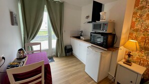 Eine kompakte Küchenzeile mit Mikrowelle, kleinem Kühlschrank und Spüle. Es gibt eine Essecke mit Stuhl und Tisch, sowie einen kleinen Schrank mit Lampe.
