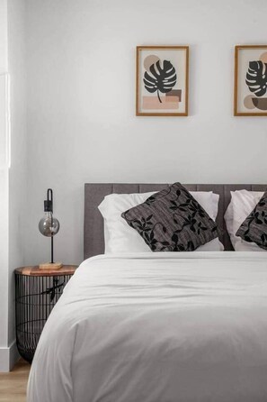 Ein ordentlich bezogenes Bett mit weißer Bettwäsche und gemusterten Kissen, eine Nachttischlampe und gerahmte Kunstwerke an der Wand.