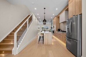A modern kitchen with a stainless steel refrigerator, wooden cabinets, a central island with stools, and a staircase with white railings. - The Olive - Luxury, 5 min 2 DT, Steps From Ctrain (Calgary)