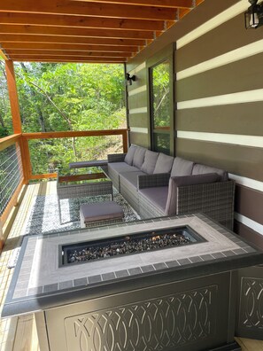 A covered outdoor seating area with a fire feature, a wicker sofa, and a view of the surrounding greenery. - Newly Remodeled Mountain Cabin - Hot Tub, Arcade & Fire Pit (Sevierville)