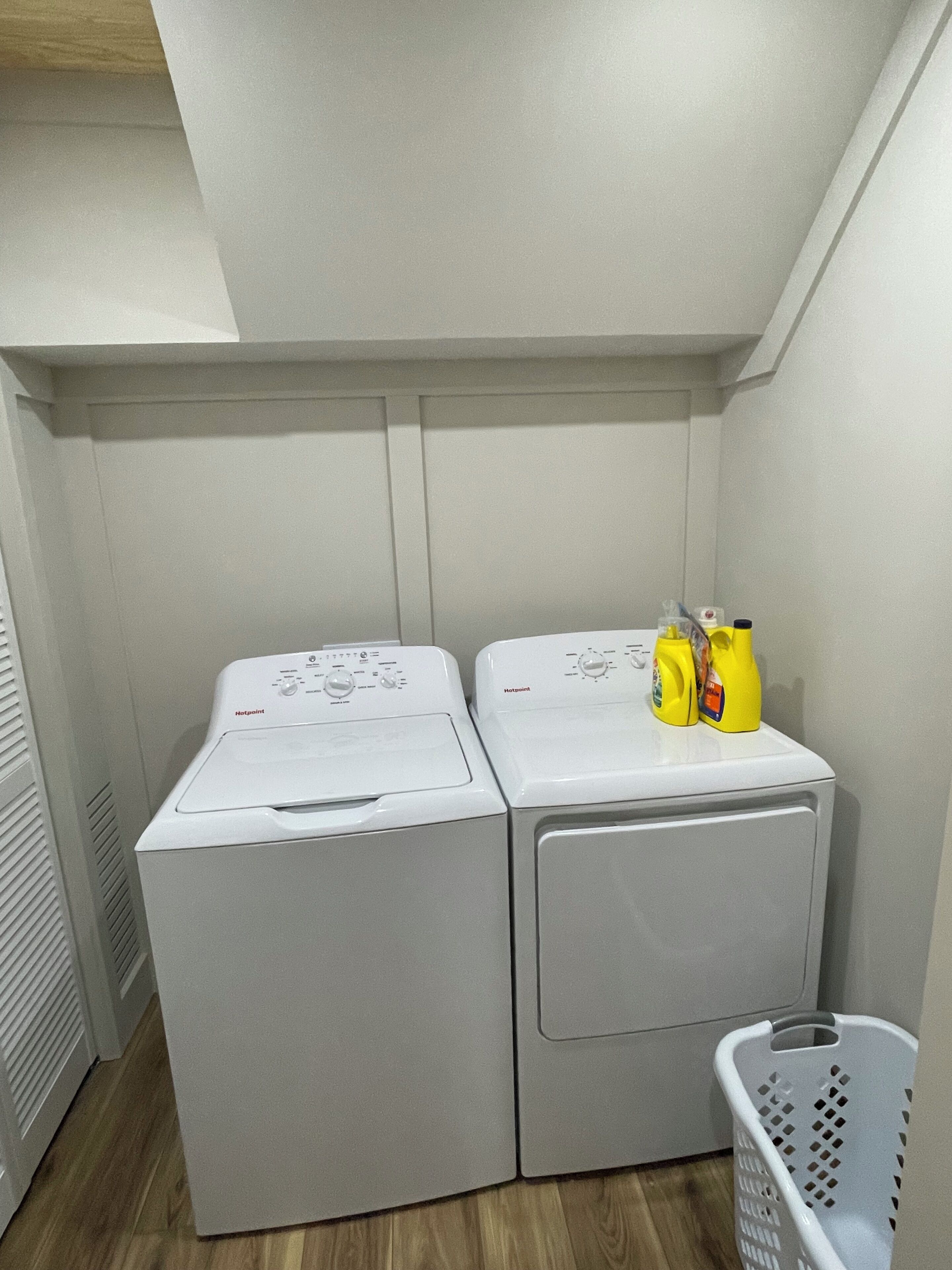 Una sala de lavandería con dos lavadoras de carga frontal blancas, una cesta de ropa blanca y dos botellas amarillas de detergente encima de la lavadora de la derecha.