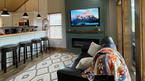 Ein Wohnzimmer mit einer schwarzen Couch, einem an einer türkisblauen Wand befestigten Fernseher, einem Deckenventilator und einer Küche im Hintergrund.