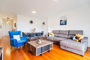 Ein modernes Wohnzimmer mit blauem Sessel, grauer Couch, Holztisch und einem Essbereich mit Tisch und Stühlen.