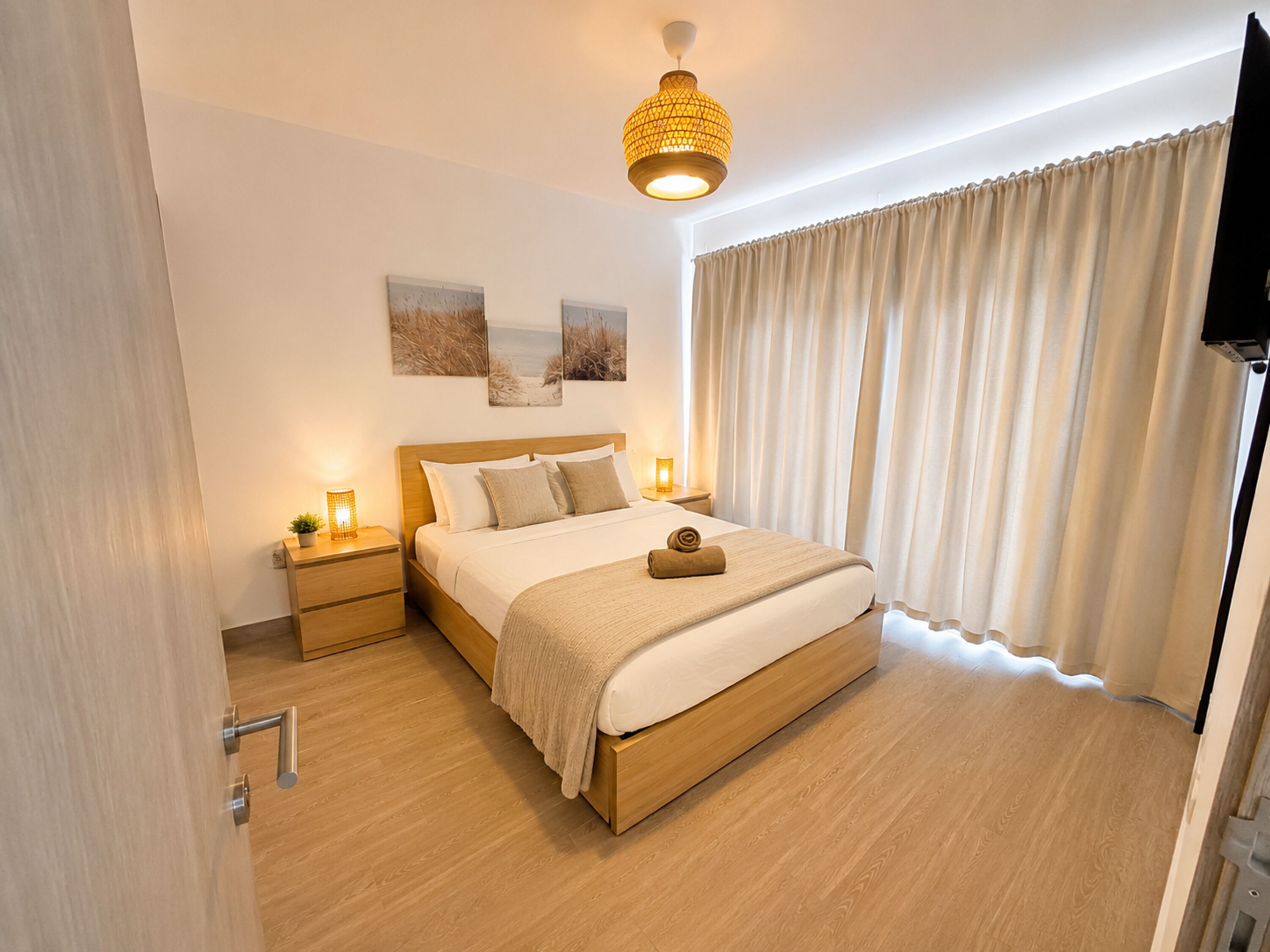 Habitación de hotel con cama de madera, dos mesitas de noche con lámparas, un ventanal con cortinas y un televisor montado en la pared.