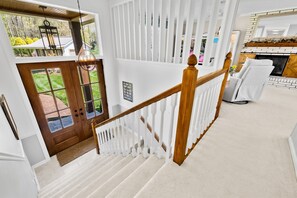 A staircase with a wooden handrail and white balusters, leading to a room with a fireplace and a view of a garden. - NEW! Hot Tub, Large Dock, Sunset Views | Sleeps 11 (Mooresville)