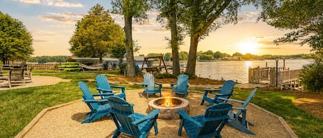 Un espace de détente circulaire, avec des fauteuils Adirondack disposés autour d’un foyer, donnant sur un lac avec un quai et une terrasse en bois.