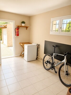 Ein kleines Zimmer mit Waschmaschine, einem Fahrrad, einem Feuerlöscher und einem Fenster mit Blick auf Grünflächen.