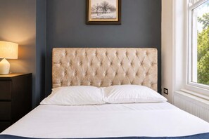 Cama com cabeceira estofada, roupa de cama branca, abajur e um quadro na parede.