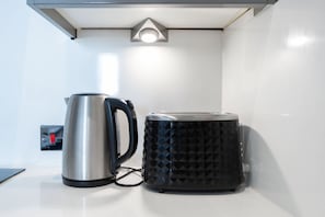 Ein Wasserkocher und eine Kaffeemaschine auf einer weißen Arbeitsplatte, darüber eine Lampe.