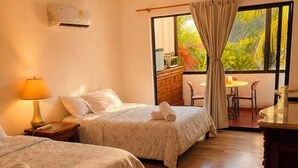 Ein Hotelzimmer mit zwei Betten, einem Deckenventilator, einer Klimaanlage, einem Esstisch mit Stühlen und Blick auf Grünflächen.