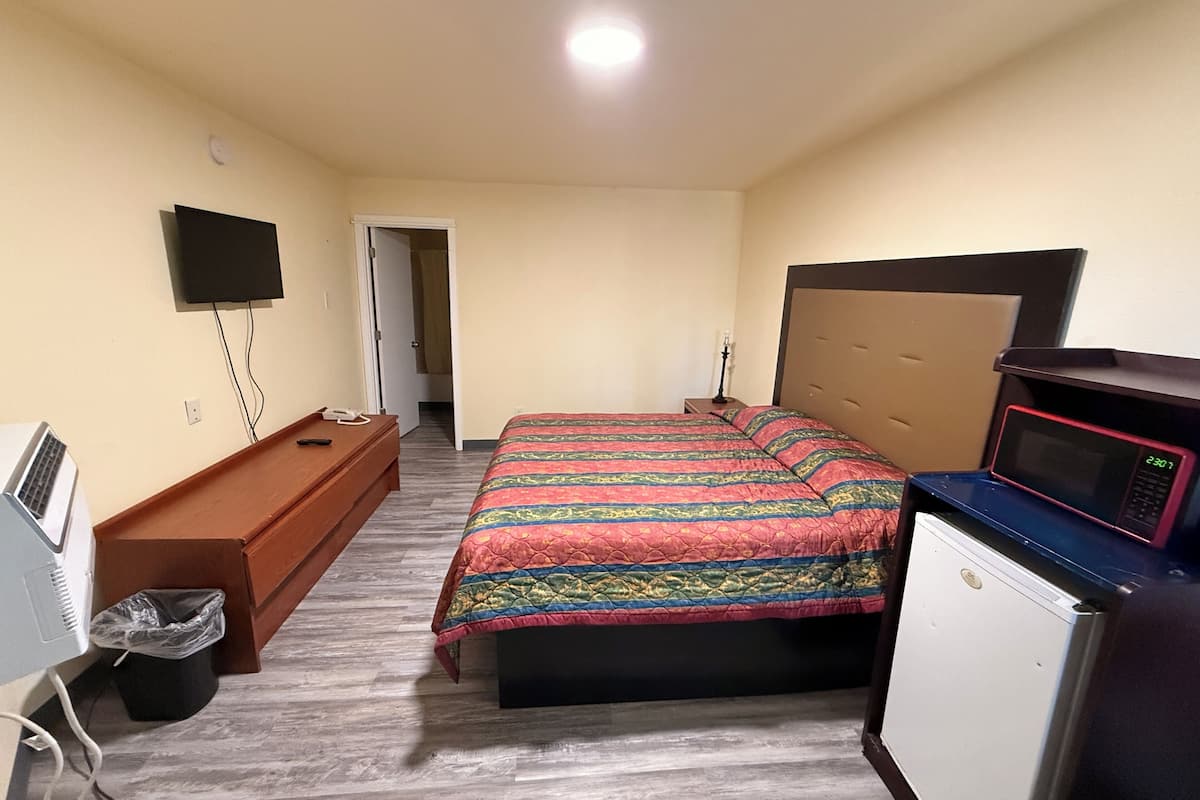 A bedroom with a bed, a TV, a desk, a microwave, and a trash bin.