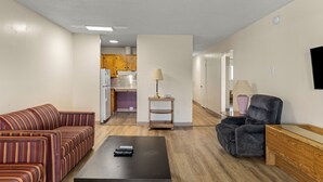 Una sala de estar con un sofá a rayas, un sillón oscuro y una mesa de centro con controles remotos. Se ve una cocina al fondo con armarios de madera y un refrigerador.